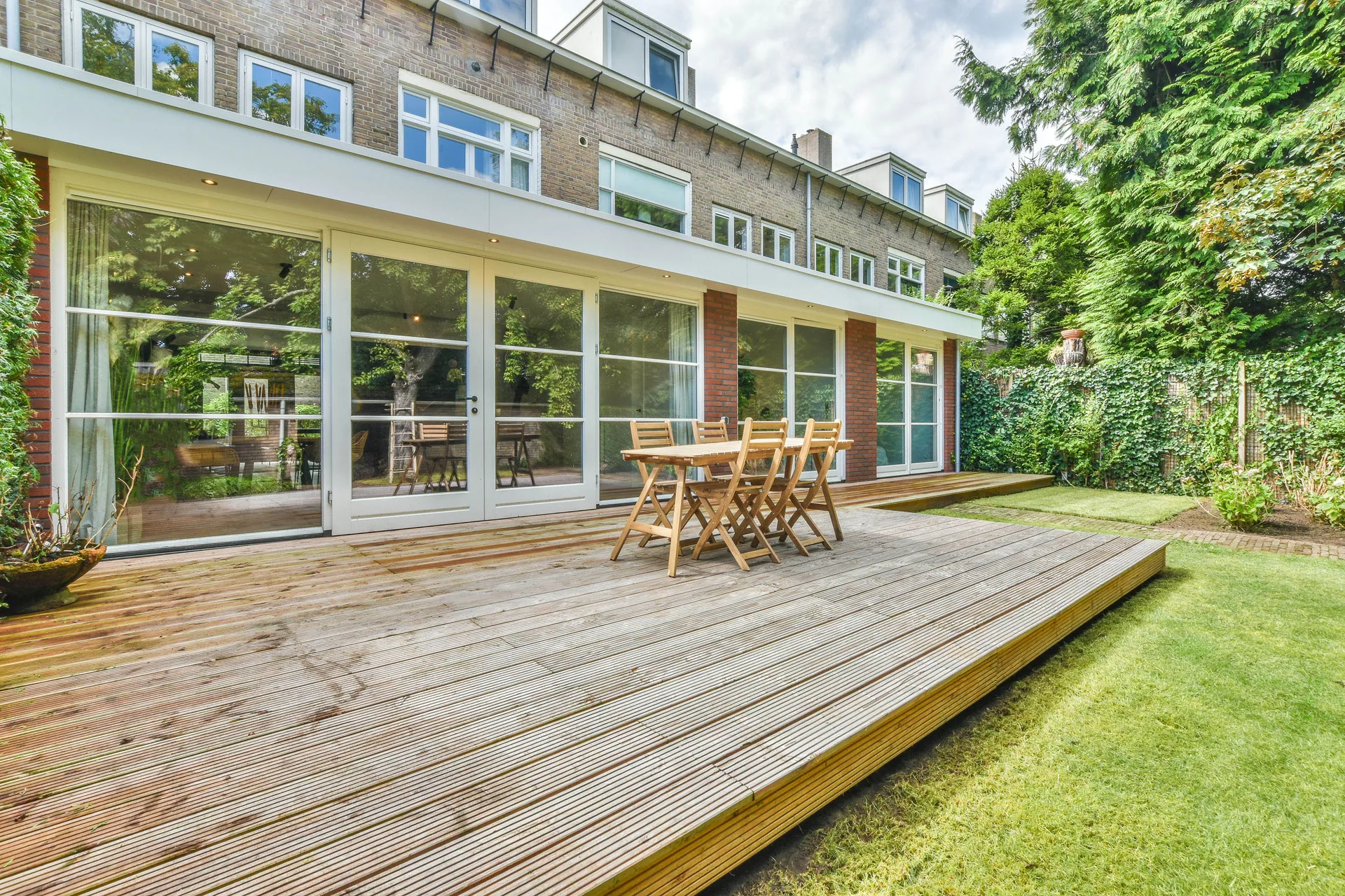 Low wooden deck with outdoor dining table and chairs behind a brick home with glass patio doors and a green lawn.