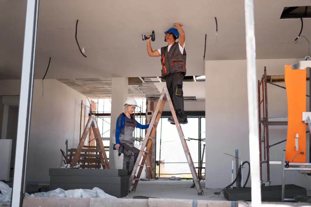 Worker on a ladder drills into a ceiling while a second worker in a hard hat stands nearby at a construction site.