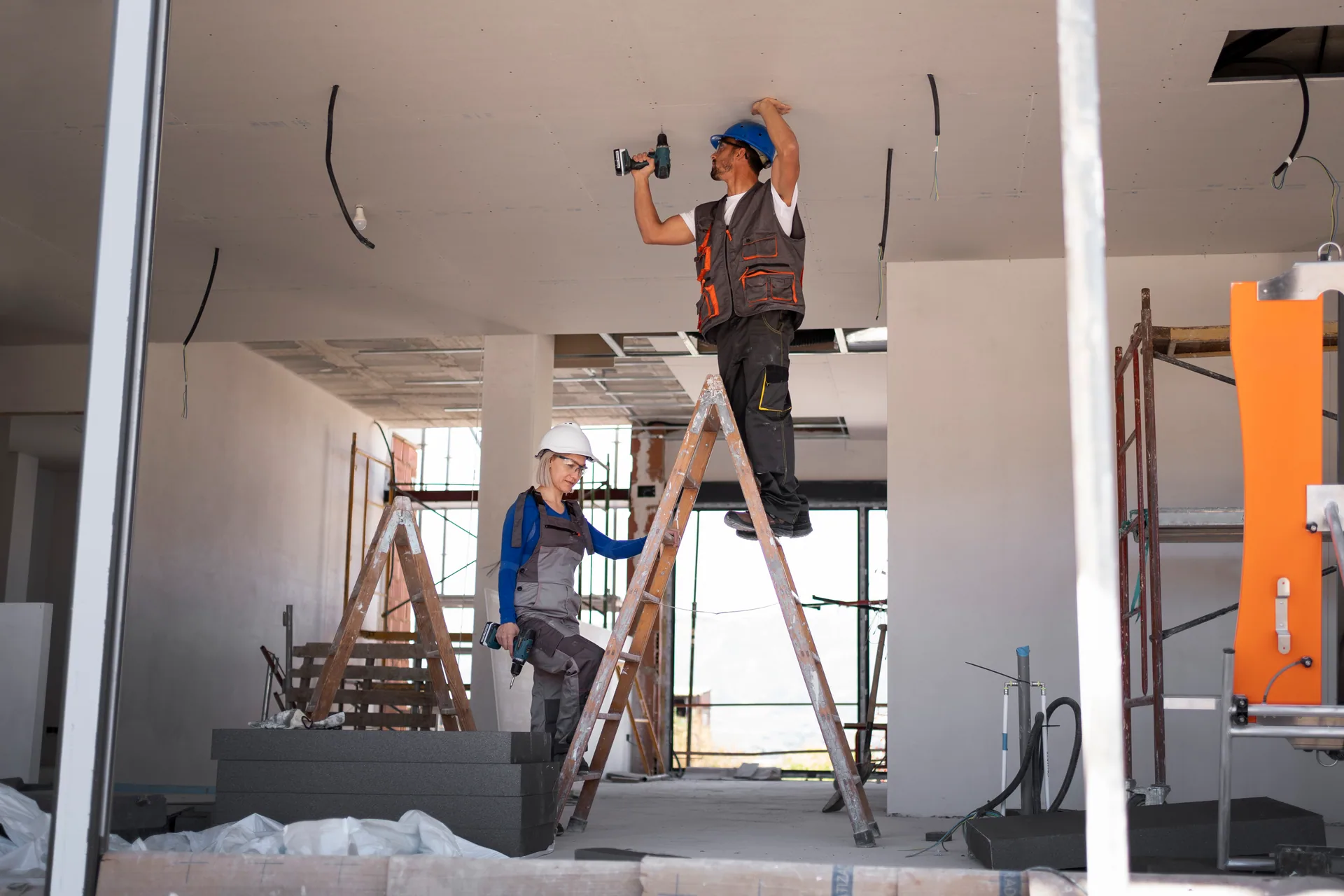 Worker on a ladder drills into a ceiling while a second worker in a hard hat stands nearby at a construction site.