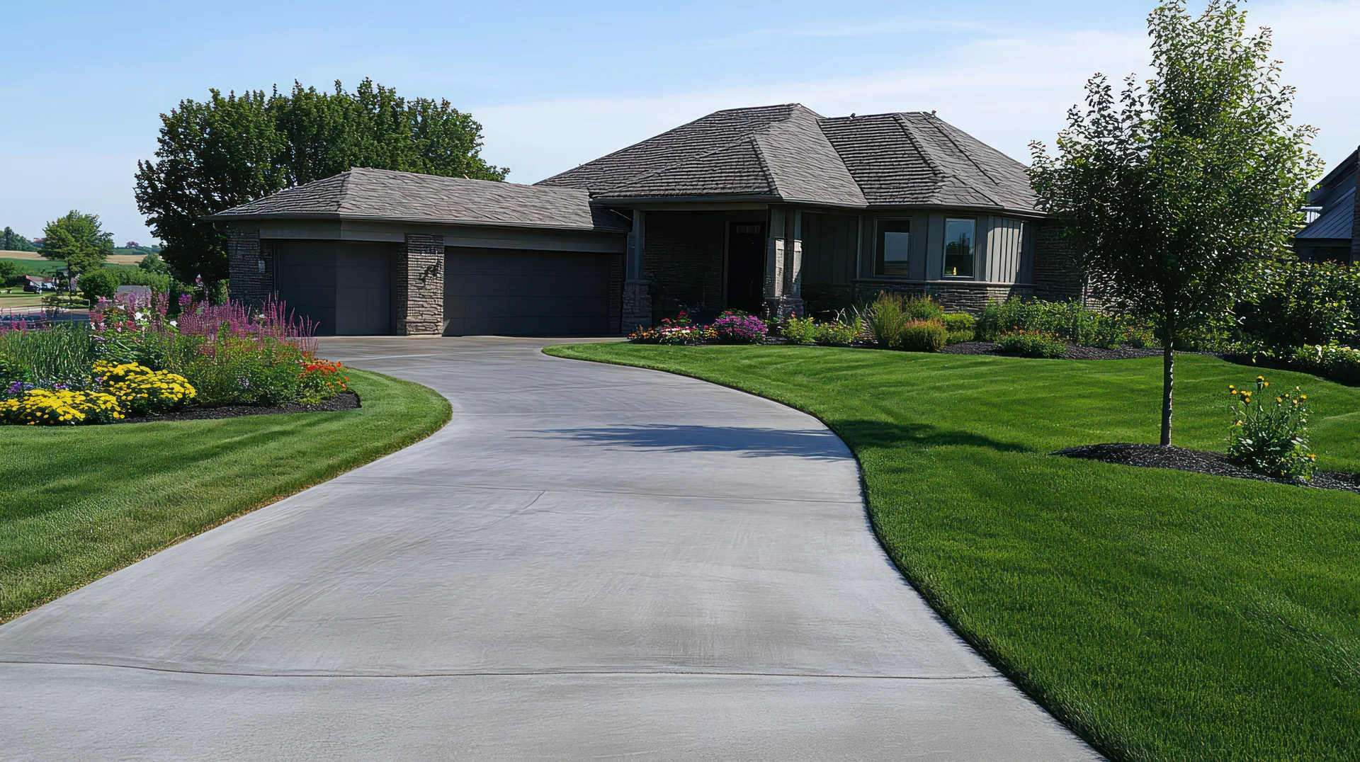 Curved poured concrete driveway leading to a ranch-style home with attached garage, green lawn, and flower beds.