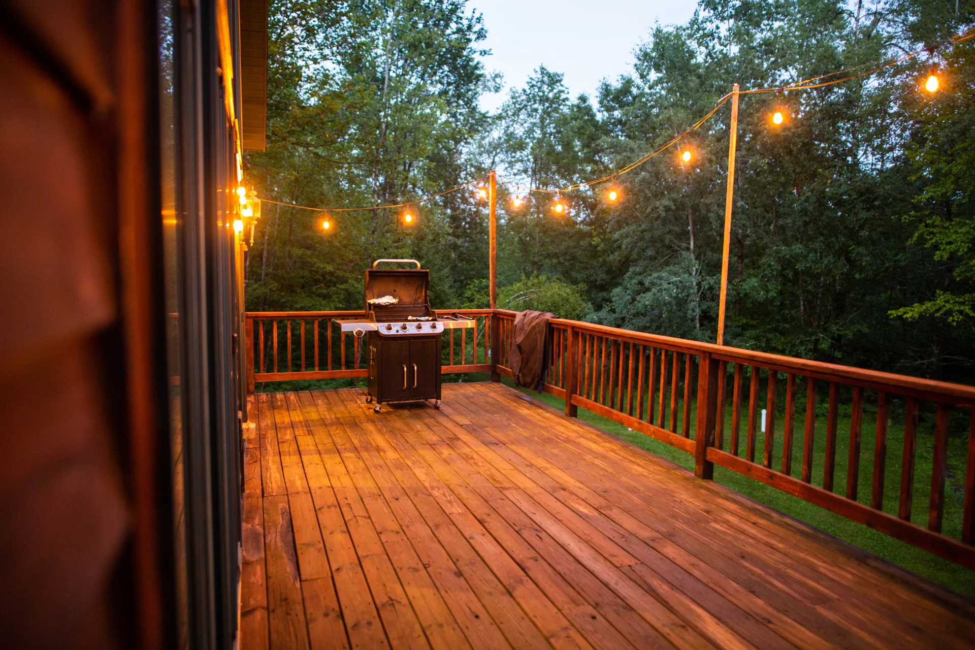 Stained wood deck at dusk with string lights overhead, a gas grill, and wooden railing surrounded by trees