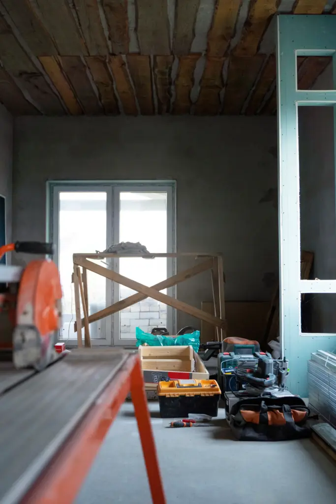 Room under renovation with a tile saw, toolboxes, power tools, drywall panels, and a wooden frame near a window.
