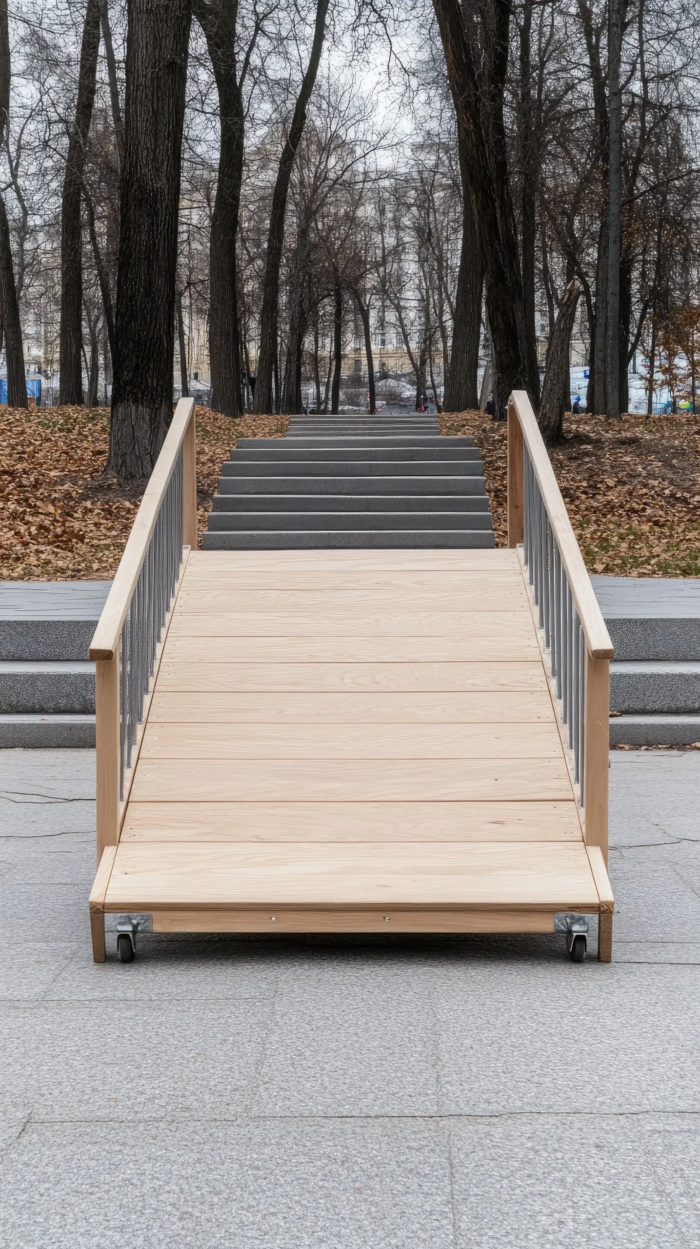 Wooden wheelchair ramp with metal balusters and wood handrails installed over stone steps in a park with bare trees.