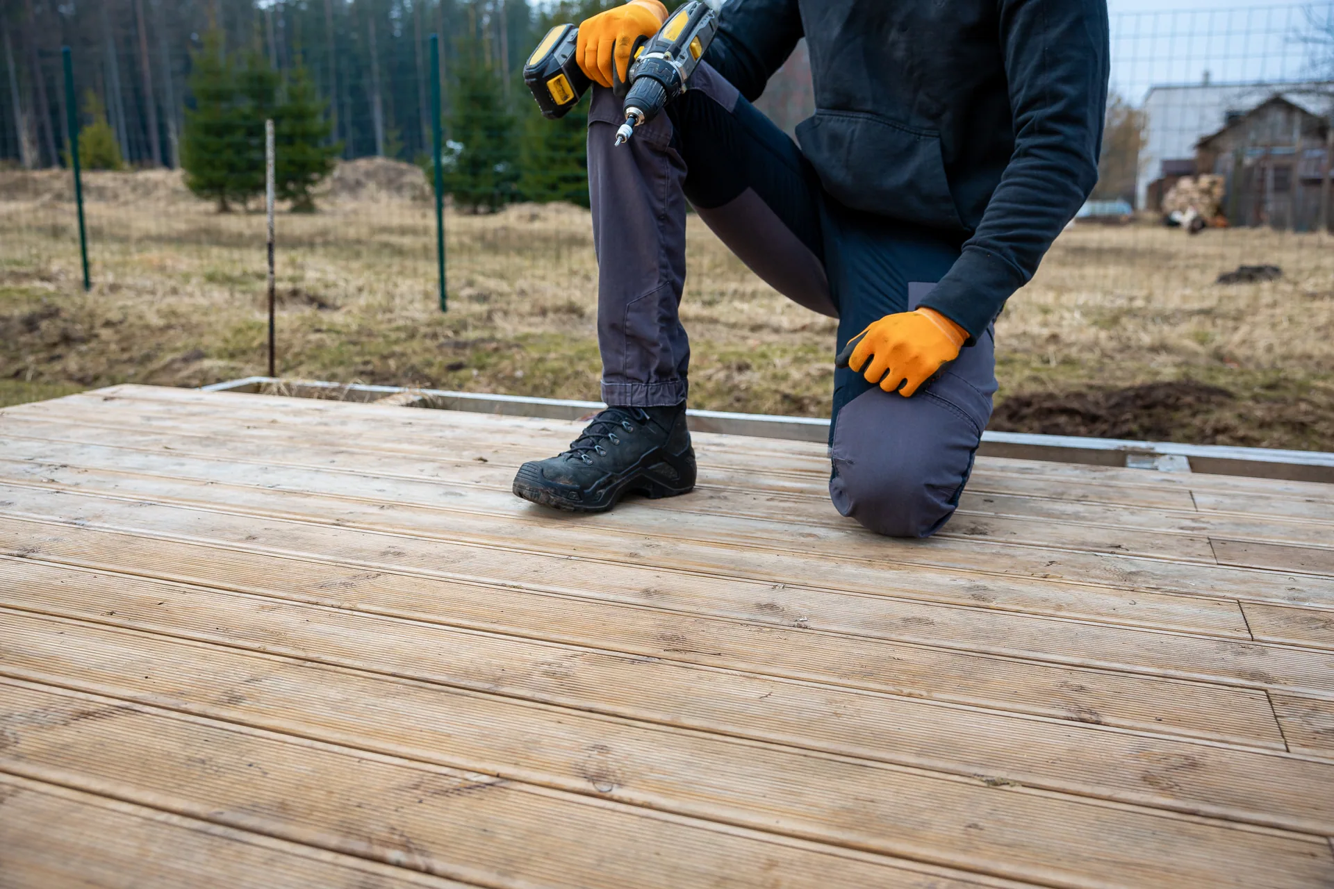 Worker kneeling on new wood deck boards holding a cordless drill, wearing orange gloves and work boots.